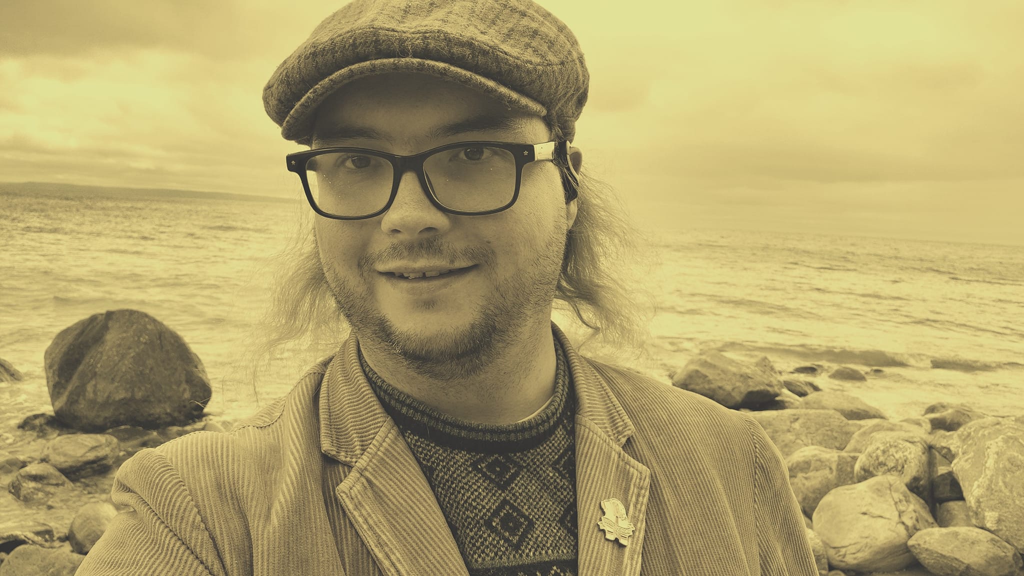 A sepia tone picture of a man wearing a corderoy jacket and flat cap standing in front of the bolder shorline of Lake Huron